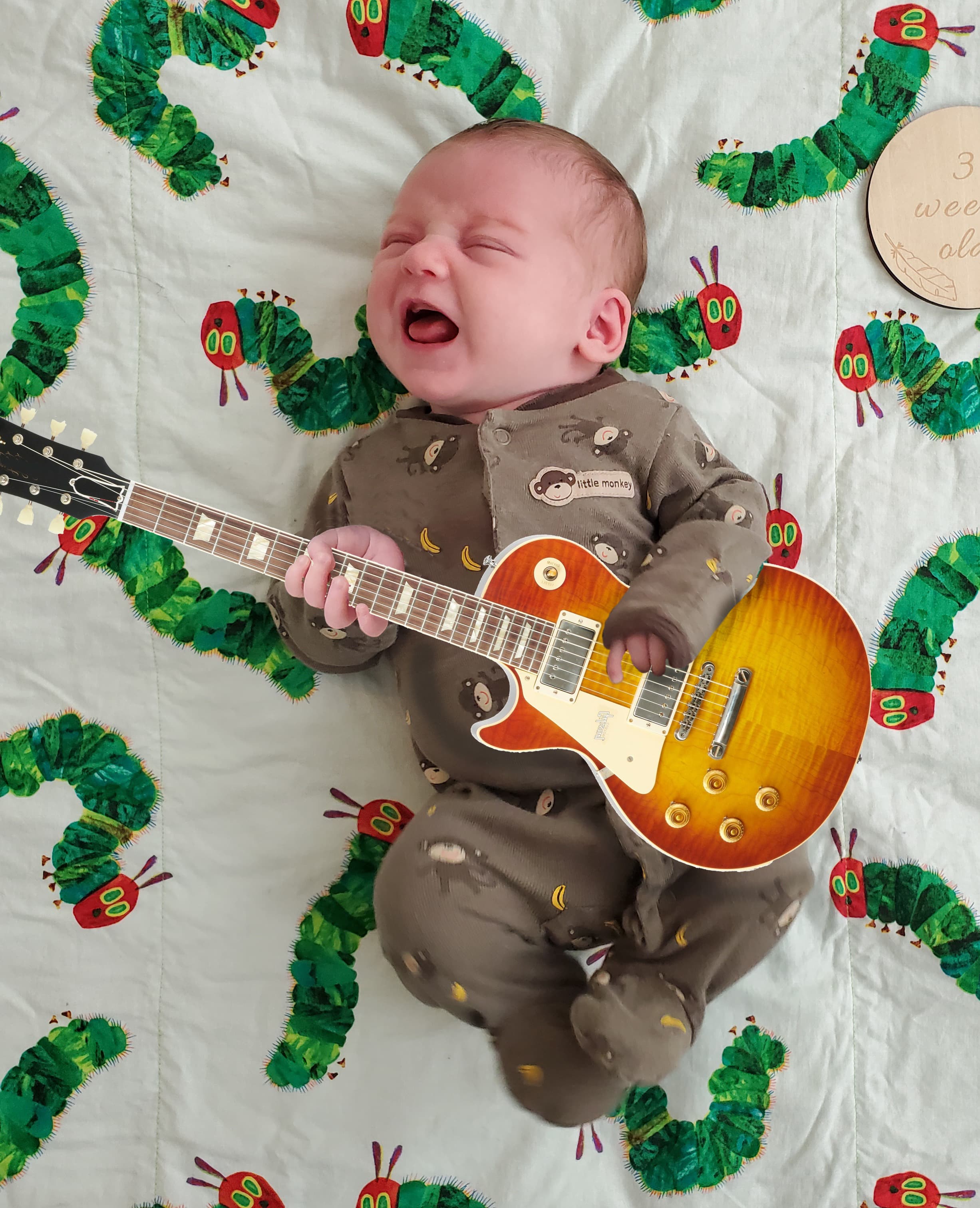 Three-week-old baby photographed on a Very Hungry Caterpillar quilt with a sunburst Les Paul guitar collaged into her arms, mid-wail